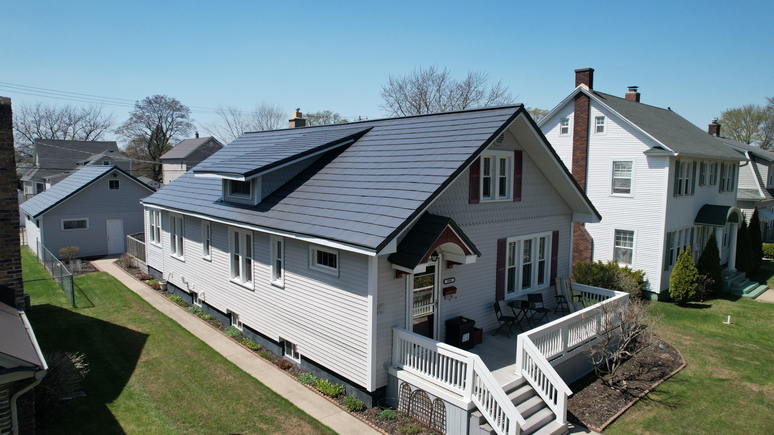 Aerial view of a historic-style home with white siding, burgundy shutters, and a charcoal gray metal roof, featuring a front dormer, covered entry, and elevated wrap-around deck with white railings, in a residential neighborhood.