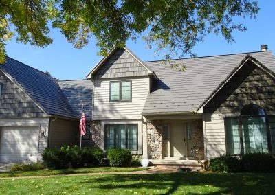 Front exterior view of a two-story craftsman-style home with gray shake siding, tan vinyl lap siding, and a charcoal gray metal roof, featuring a natural stone accent entry