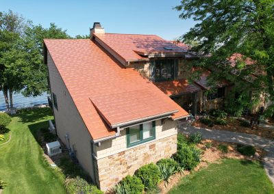 Aerial view of a multi-level lakefront home with natural stone and shingle siding, a copper-toned metal roof
