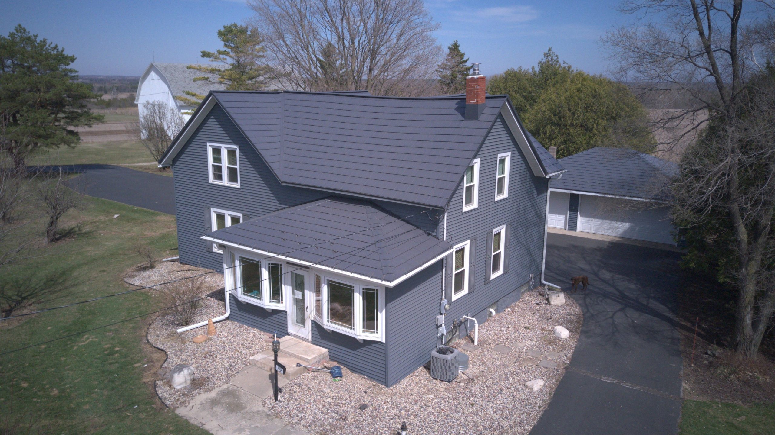 Aerial view of a two-story farmhouse with dark navy siding and a charcoal gray metal roof, featuring a sunroom addition, brick chimney, and a detached garage, surrounded by gravel landscaping on a rural property.