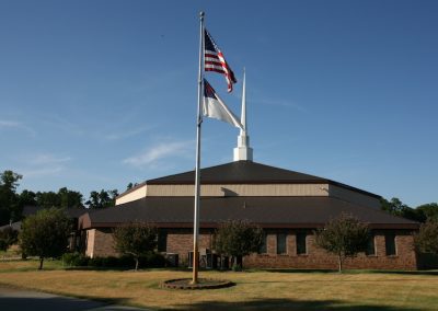 Exterior view of a single-story brick church with a dark brown metal roof and a white steeple, fronted by a flagpole flying the American flag and a Christian flag, with a manicured lawn and ornamental trees under a clear blue sky.